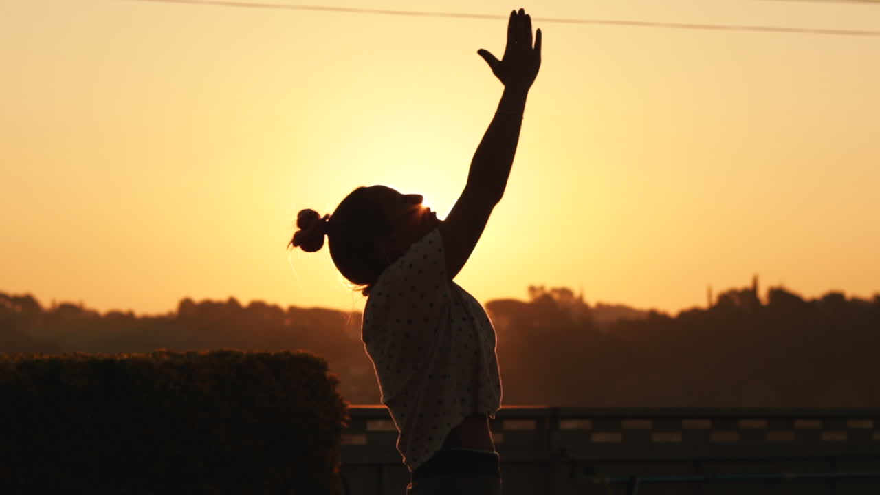 Silhouette of young woman relaxing from yoga pose at sunrise in slow motion with a strong sun backlight. The sun shines between the silhouette with a beautiful flare