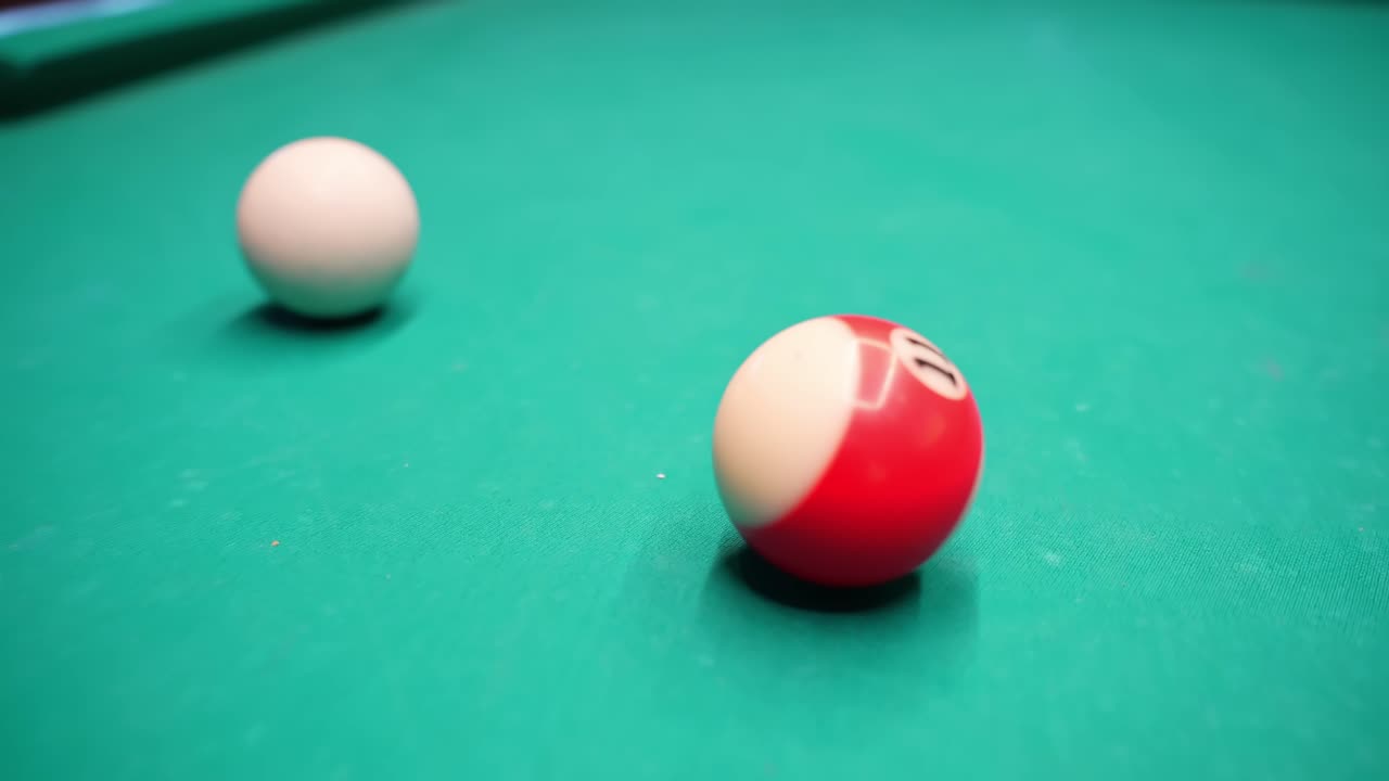 Close-up on a green pool table showing the white cue ball and a red striped ball in shallow focus, cinematic indoor bar game footage with clean copy space