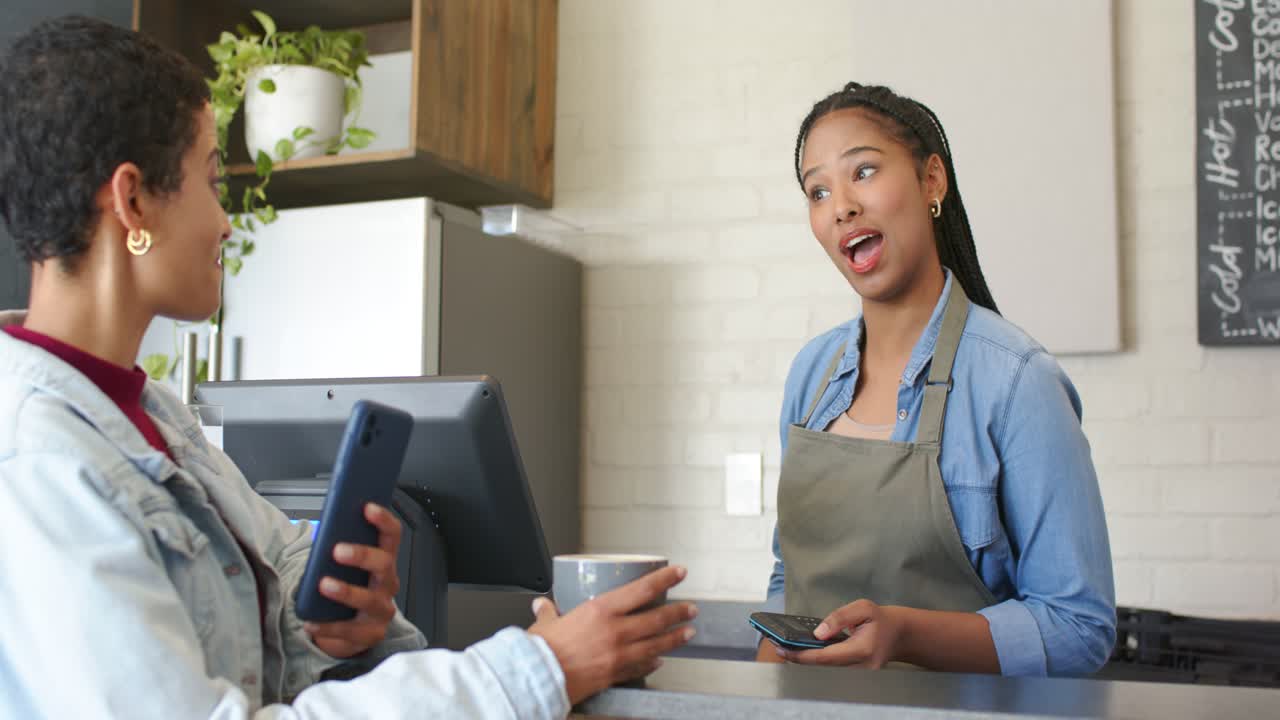 female customer tapping smartphone completing payment while barista holding reader, smiling