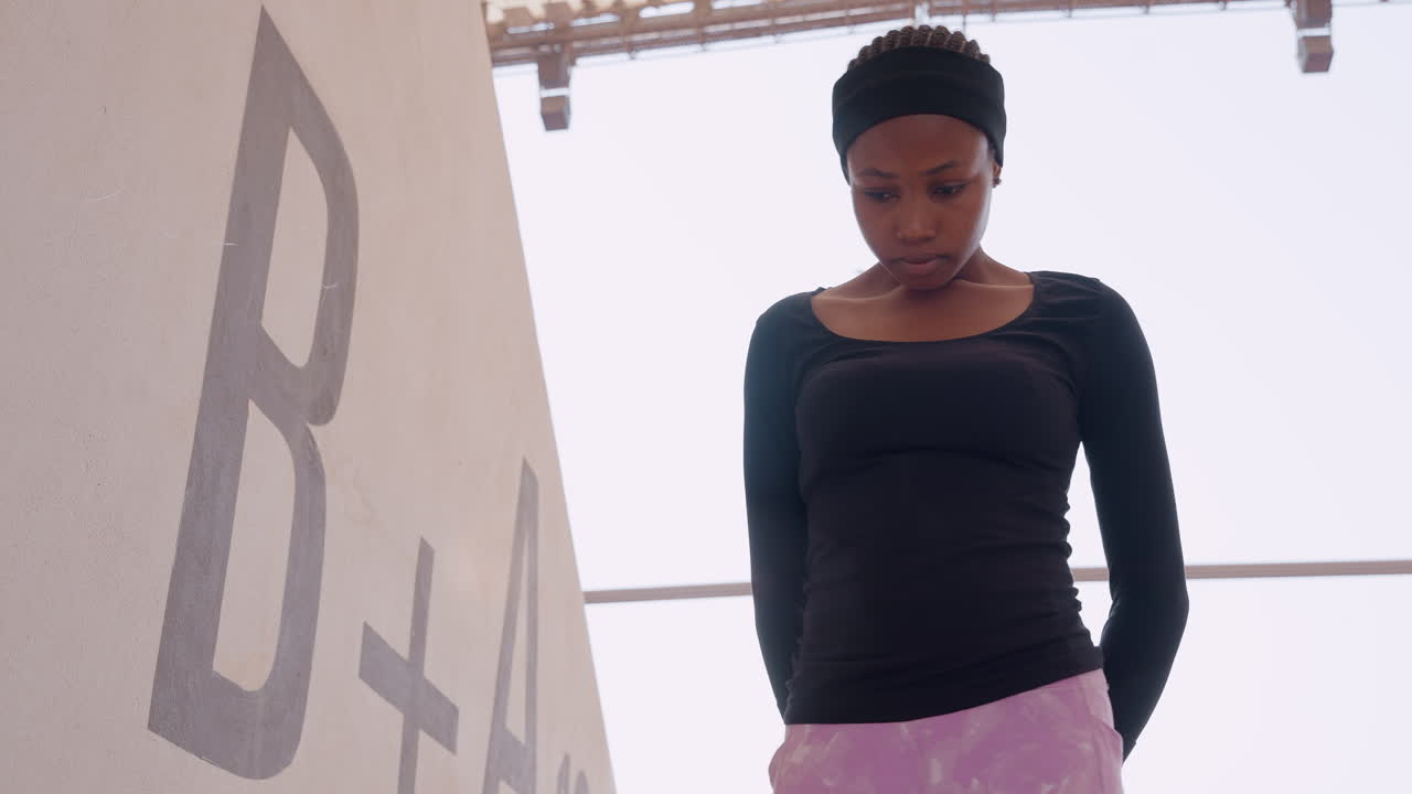 black woman standing by concrete wall reflecting downward near B marking, wearing headband and casual top, soft daylight, urban architecture, contemplative mood, cinematic closeup