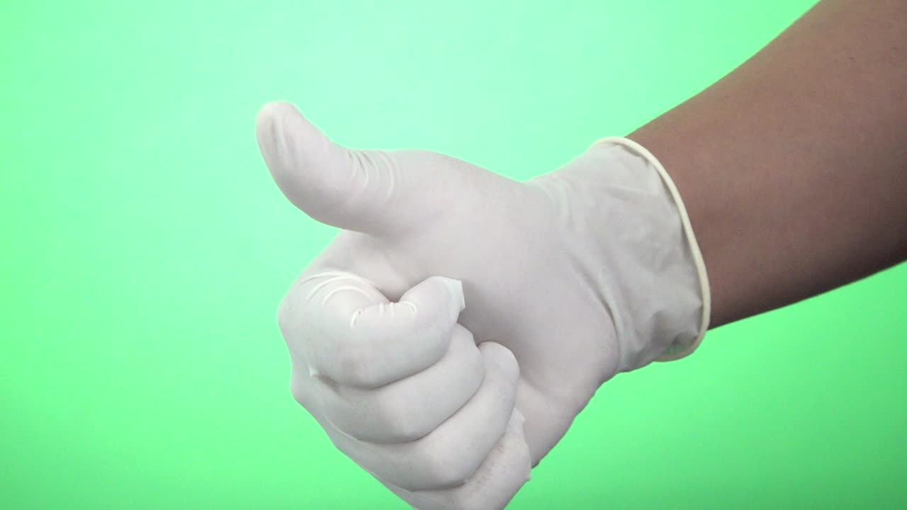 Close-up shot of left and right hands of a woman alternating in frame wearing white latex gloves and making a thumbs-up gesture on a green screen background
