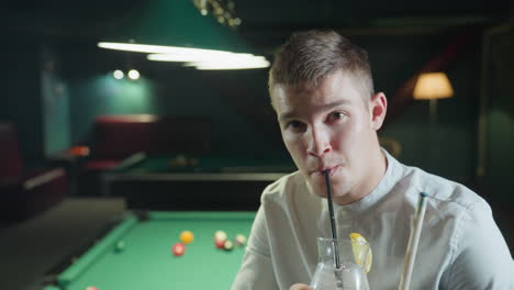 close-up of billiard player wearing white shirt, sitting on green pool table, holding cue stick and glass of lemon drink with black straw. he raises the glass and takes a sip, with a slight smile