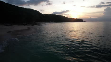 Drone-flying-low-over-sea-surface-with-sunlight-reflection,-Tropical-beach-Sunset