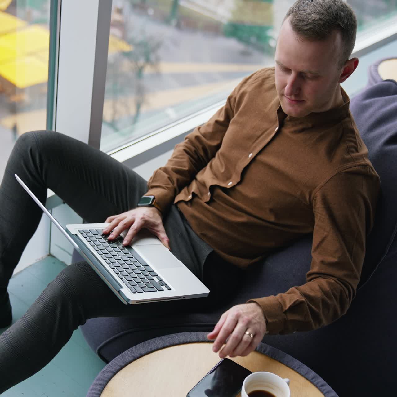 Handsome businessman in office by the window. Young man in brown shirt and trousers sitting on a comfortable ottoman and working on a laptop. City view background