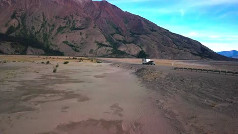 Sensational-travel-scene-of-truck-pulling-tag-along-RV-camper-trailer-in-remote-rural-Yukon-wilderness-highway-by-colorful,-imposing-and-impressive-Sheep-mountain-by-Kluane-lake,-aerial-approach