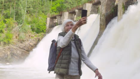 anciana emocionada con el pelo gris parada frente al aliviadero de la presa y llamando por video a alguien por teléfono móvil