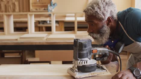 African-american-male-carpenter-using-an-electric-grinder-to-grind-wooden-plank-at-a-carpentry-shop