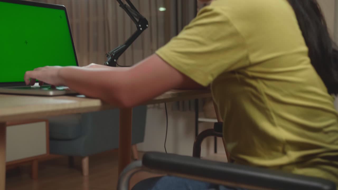 Back View Of Upset Asian Kid Girl Sitting In A Wheelchair While Using Laptop Computer With Mock Up Green Screen At Home