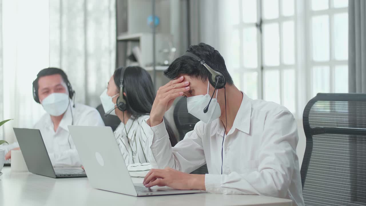 A Man Of Three Asian Call Center Agents Wearing Headset And Mask Is Tired Because Two Of His Colleagues Are Talking During Working At The Office