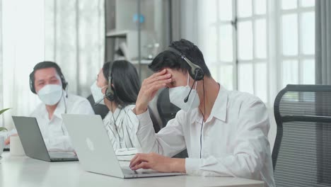a man of three asian call center agents wearing headset and mask is tired because two of his colleagues are talking during working at the office