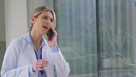 Female-Doctor-Wearing-White-Coat-Standing-In-Hospital-Corridor-Talking-On-Mobile-Phone