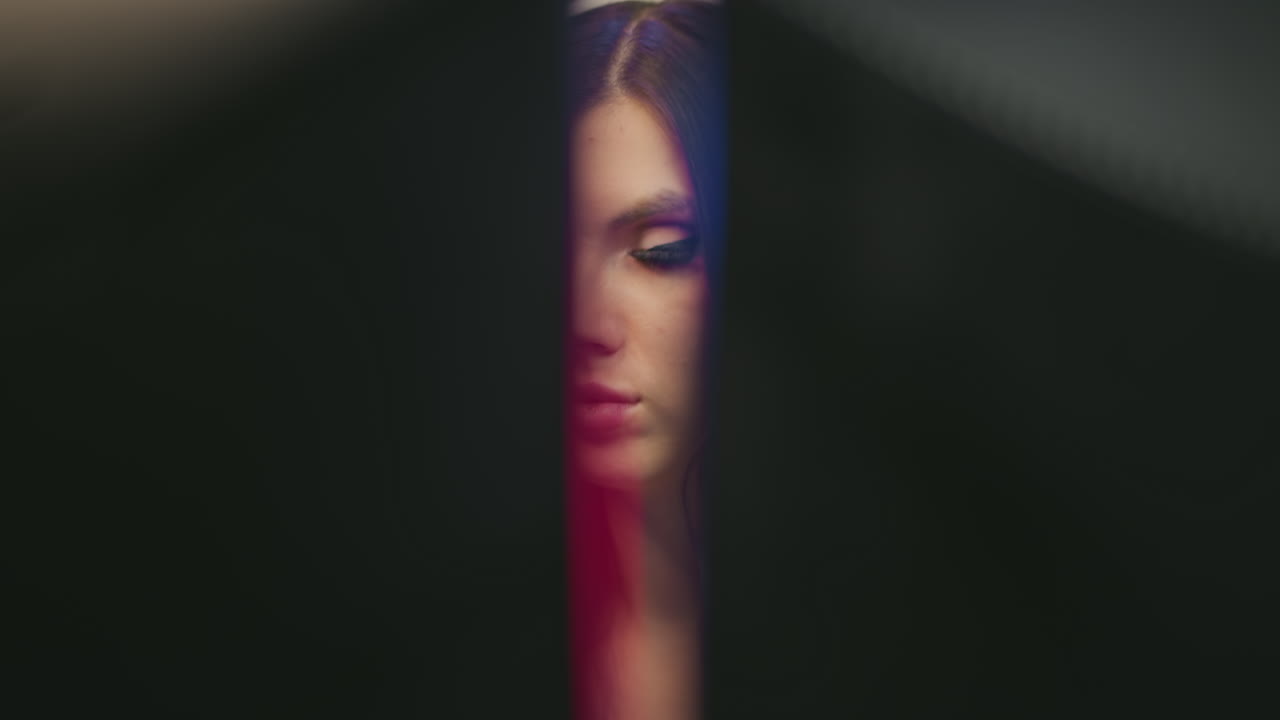 female wearing white headphones peering intensely through gap between dual desktop monitors in neon lit purple studio environment focusing on computer screen during late night creative gaming session