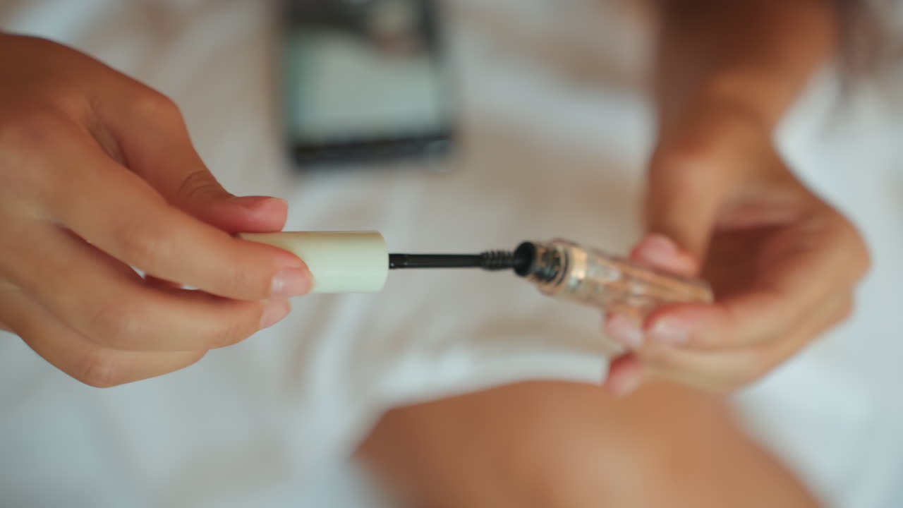 Arreglo personal íntimo por la mañana. Mujer aplica máscara de pestañas pacíficamente en el interior. Primer plano de la mano con la máscara de pestañas cerca de un smartphone. Mujer aplica cuidadosamente la máscara de pestañas en un tranquilo ambiente matutino de hotel.