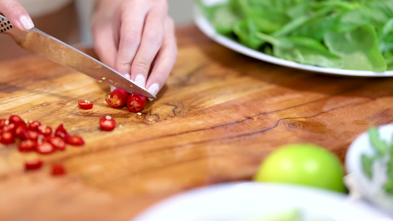 Close-up of hands slicing red chili on a wooden board with fresh greens nearby