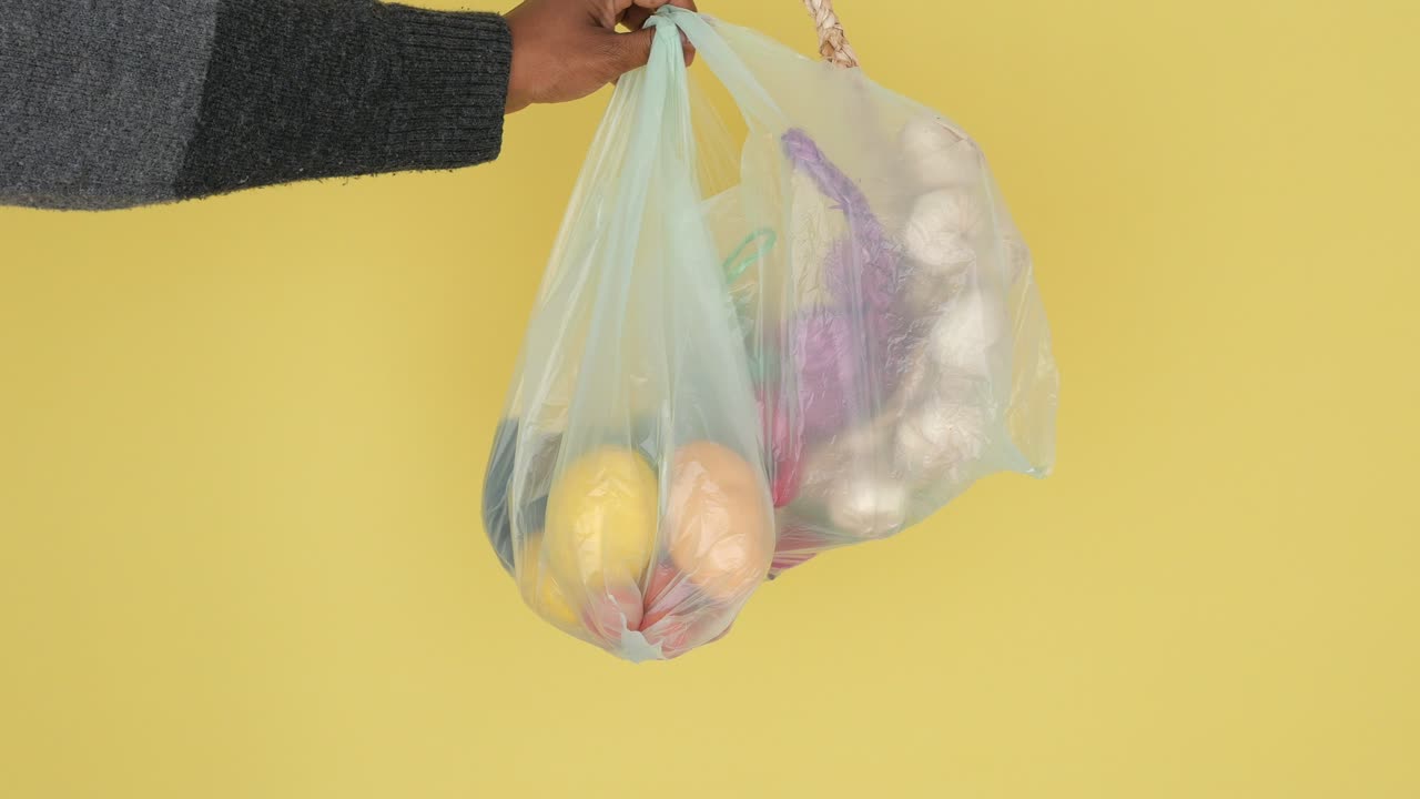Hand carrying a plastic bag filled with fresh fruits and vegetables on a yellow background