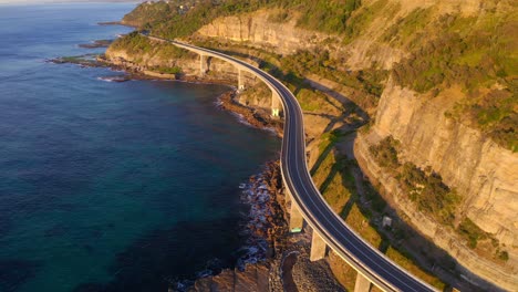 sea cliff bridge section of the grand pacific drive around steep sandstone cliff at the edge of pacific coast during sunset - aerial drone shot