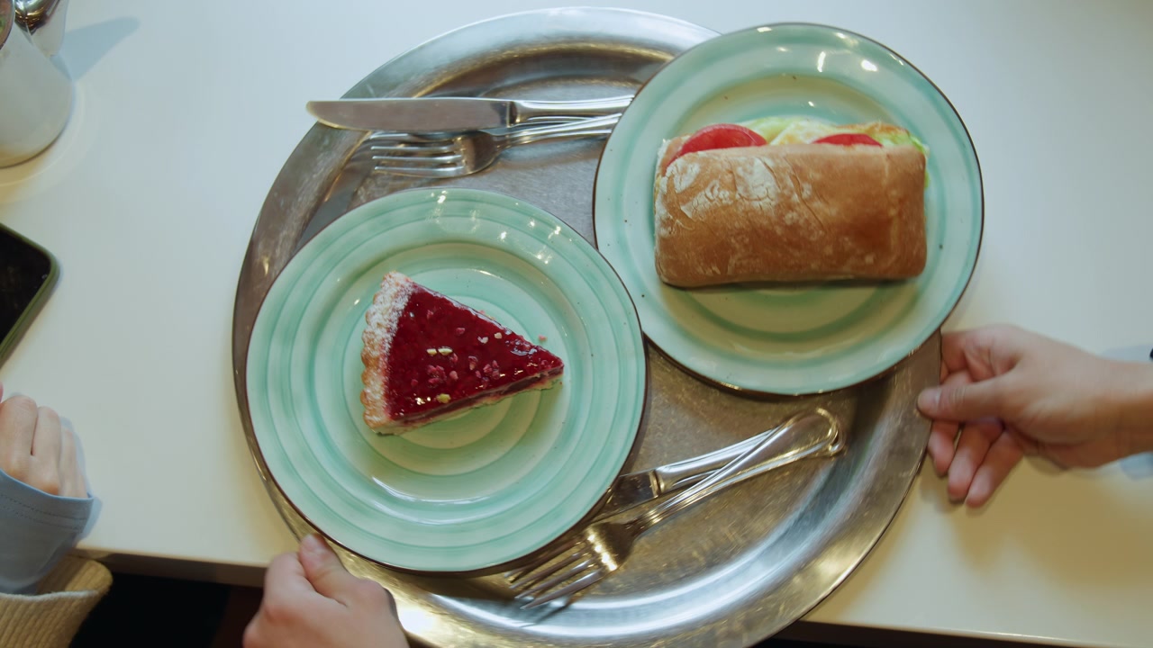 Nærbillede af sandwich og tærte på tallerkener med bestik, mens hænder rækker ud efter bakken. Scenen viser madvarer pænt arrangeret i blød belysning, der fremhæver tekstur, detaljer og komposition.