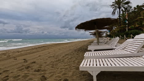 Toma-4k-De-Una-Playa-Vacía-Con-Tumbonas-Y-Sombrillas-De-Paja-En-Marbella,-España