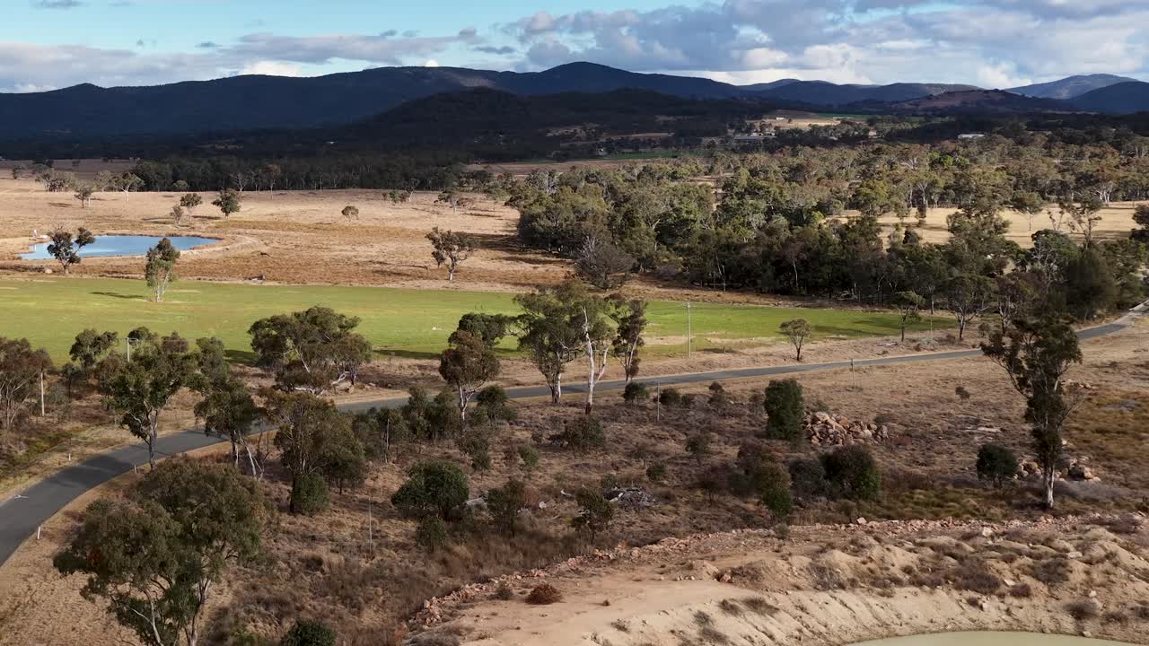 Aerial camera slowly pans across a rural landscape with farmland, trees, a dam, and distant mountains under partly cloudy daylight in Stanthorpe, Queensland