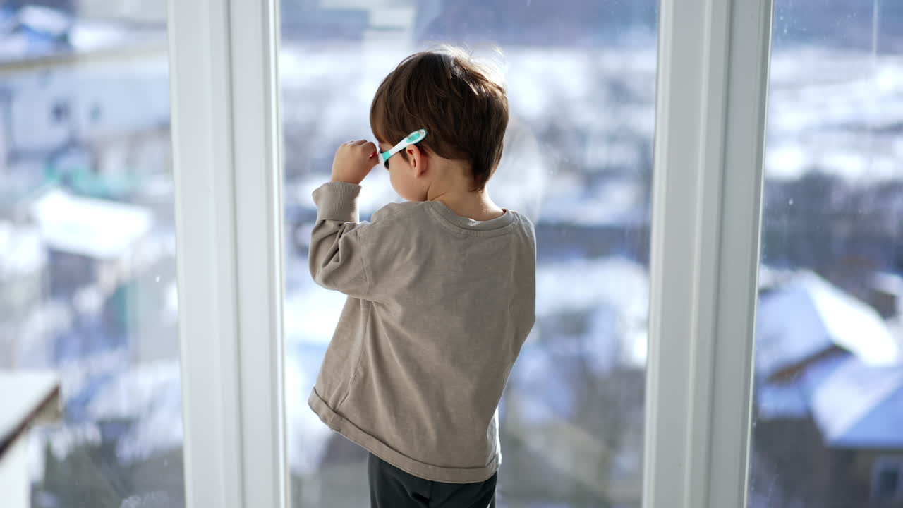 Baby boy stands at the window wearing the sunglasses. Kid turns to camera and points at the window.