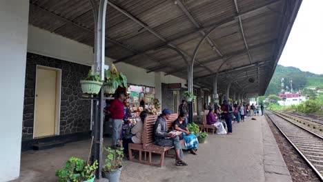 a diverse group of locals sitting on a platform bench at haputale station, sri lanka, waiting for a train amidst scenic hills.