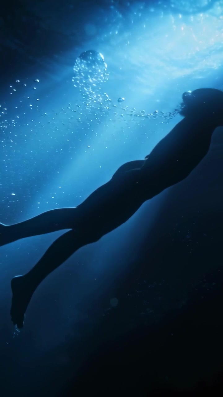 Vertical video: Gliding swimmer in swimwear exhaling bubbles moving to top seeking air in sunbeam