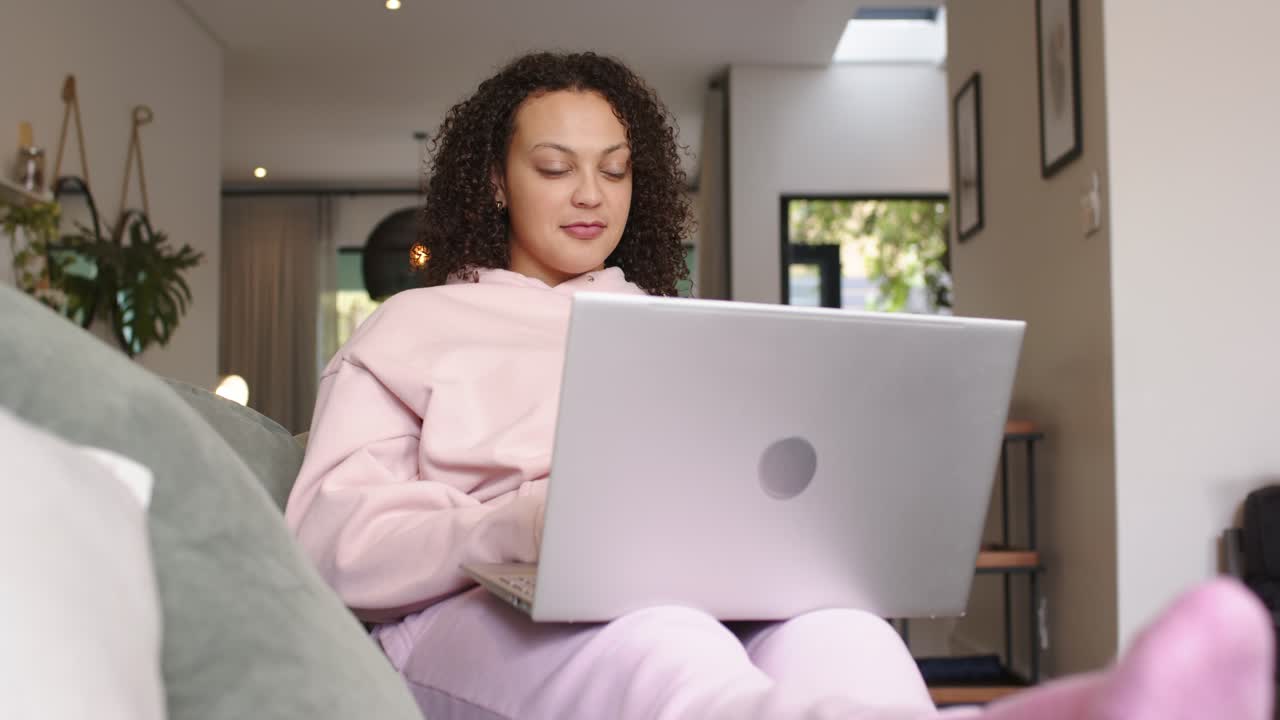 Mulher afro-americana na casa dos 20 anos abrindo um laptop prateado no sofá, sorrindo, digitando e trabalhando na sala de estar.