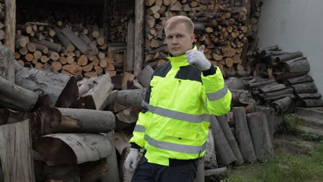 Lumberjack-in-reflective-jacket.-Man-woodcutter-show-ok-sign.-Sawn-logs,-firewood-background