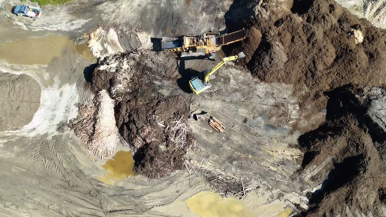 Drone footage captures an excavator handling mulch piles in a dirt field under bright daylight, showcasing industrial processing