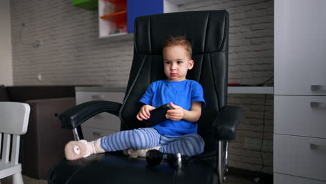 serious baby boy in blue t-shirt sits in a big black chair. cute kid holding phone in hands and then puts it to the ear.