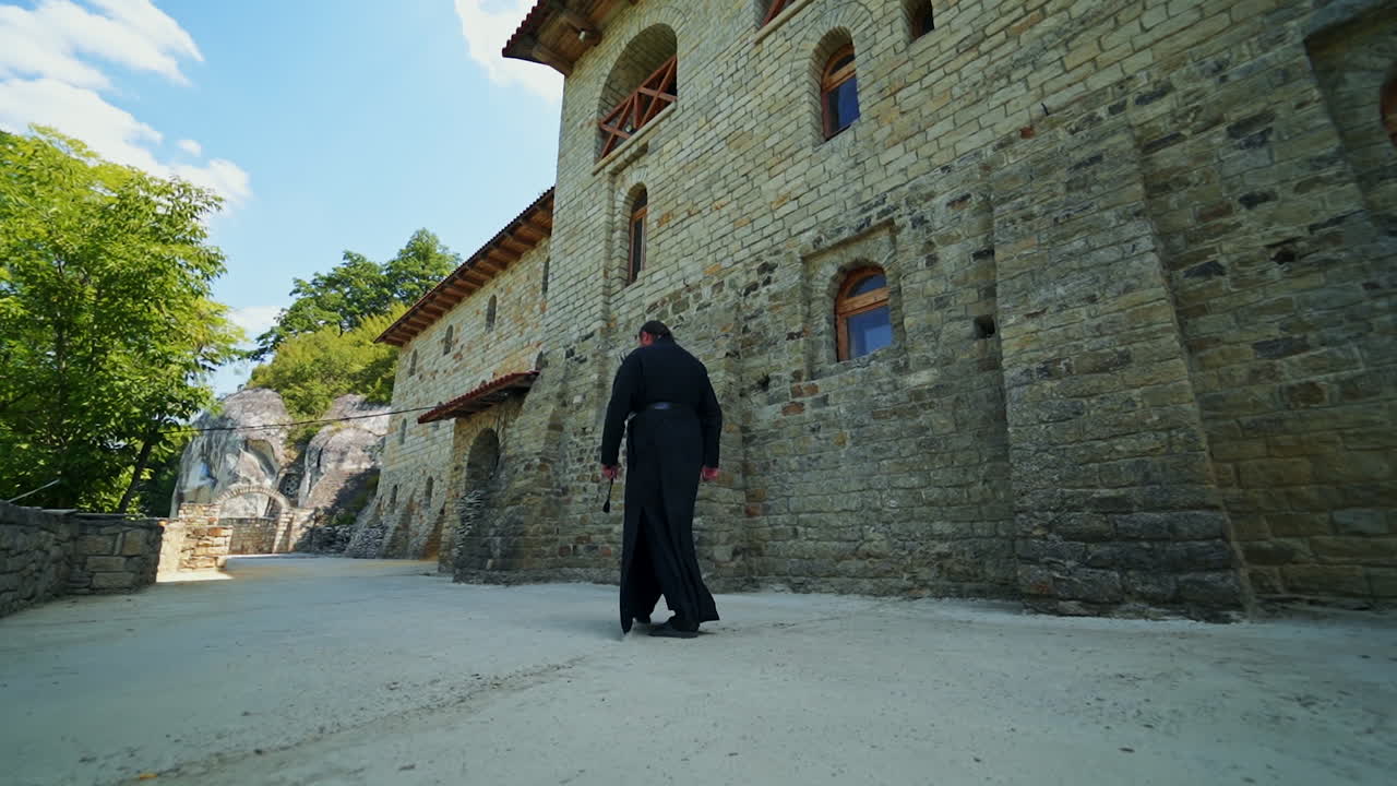 Sacerdote de vestes pretas caminhando ao longo dos muros de pedra do mosteiro. Religioso da igreja ortodoxa. Vista de baixo para cima