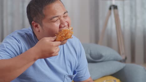 close up of a fat asian man eating fast food having fried chicken then drinking a glass of coca cola on a sofa in the living room at home