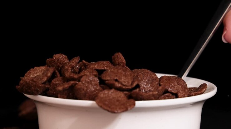 A spoon drops chocolate cereal into a white bowl against a black background, highlighting the crunchy texture and rich color