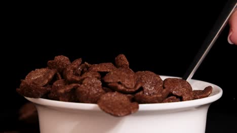 a spoon drops chocolate cereal into a white bowl against a black background, highlighting the crunchy texture and rich color