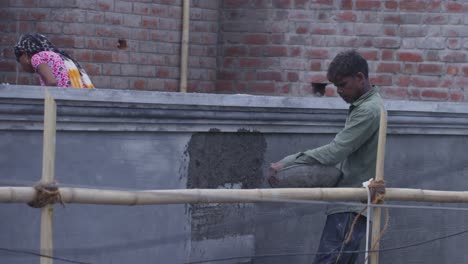 a slow motion handheld long wide shot of an indian woman wearing pink kurta and scarf covered on head and a man wearing faded olive green shirt, working at a construction site, applying cement on wall