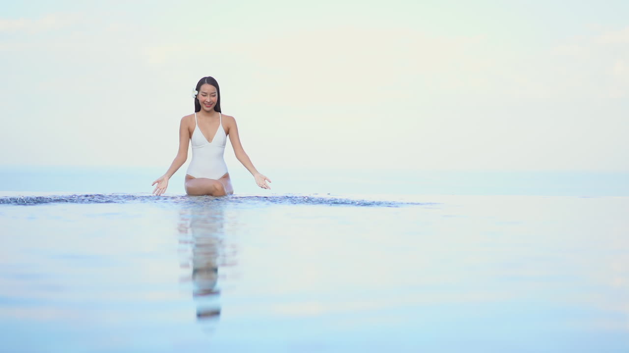 인피니티 풀 가장자리에 앉아 머리에 꽃을 꽂은 아시아 소녀가 물놀이를 합니다.