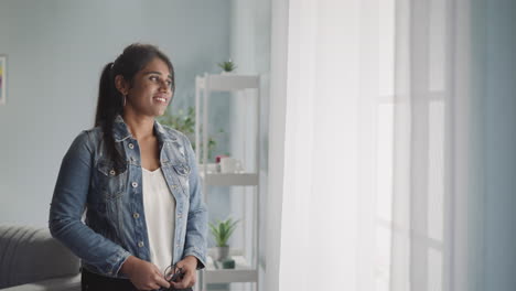 pensive indian woman in denim jacket looks out bright window