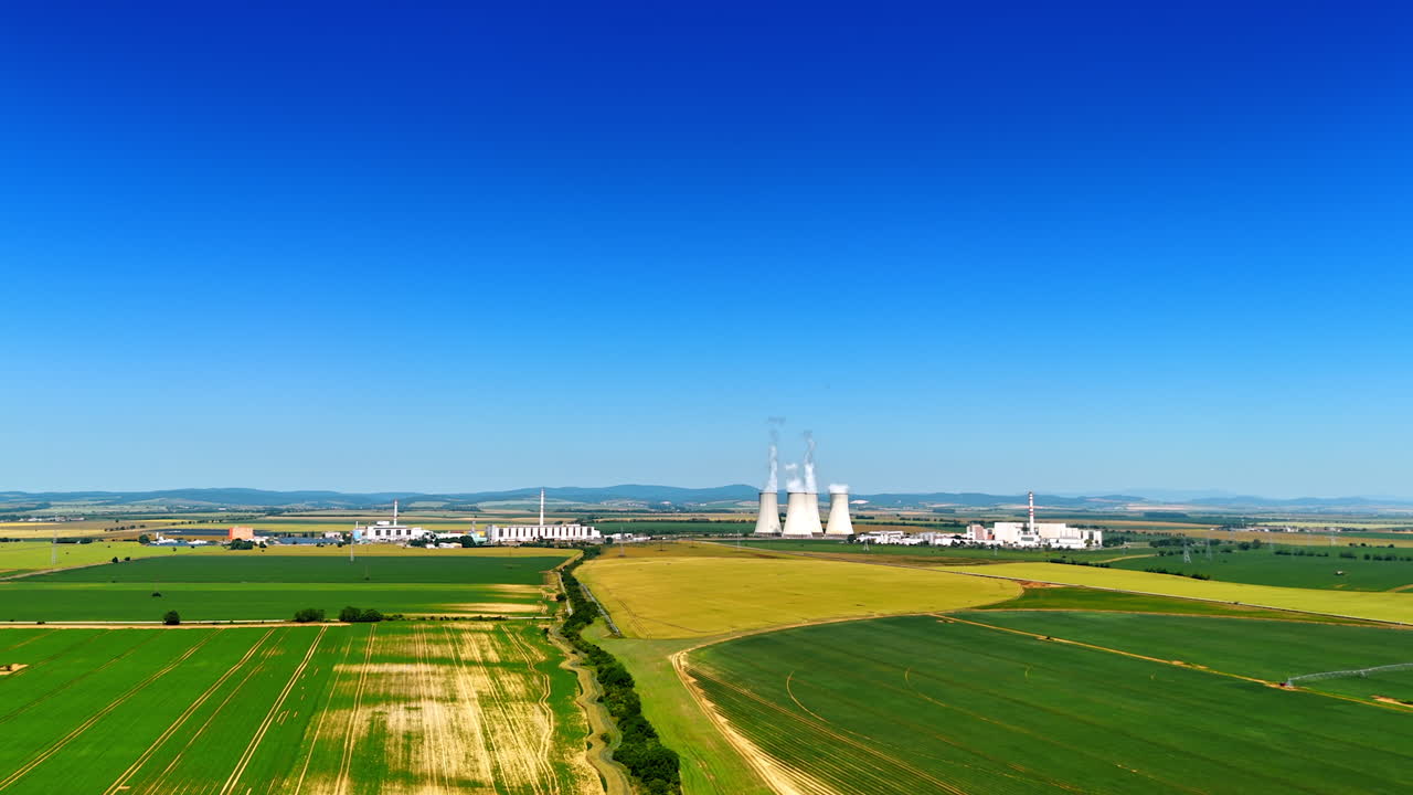 Flight above the green countryside on sunny day. Aerial perspective on the large factory producing white smoke