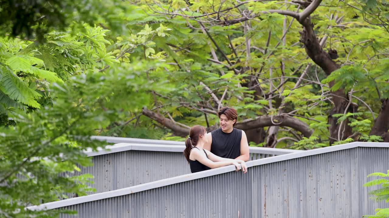 黒いスポーツウェアを着た健康的な若いカップルが、公園の橋の手すりに寄りかかって話しています。