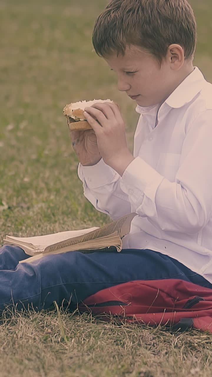 pupil in white shirt and jeans enjoys burger sitting on grass lawn with textbook on knees close view slow motion