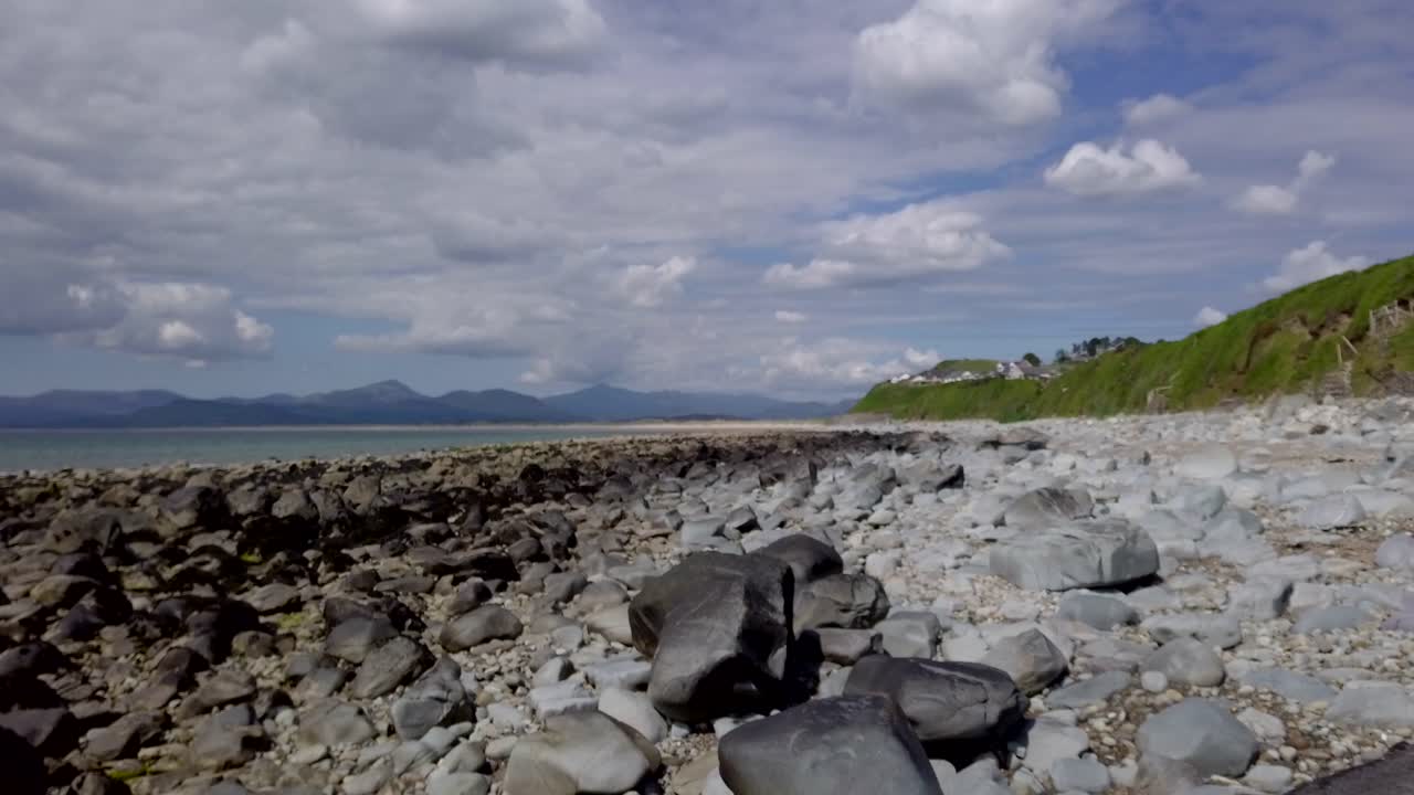 A slow Right to Left smooth Drone track across a rocky beach to darker rocks with distant mountain in the distance in Wales in the UK - 5 second version