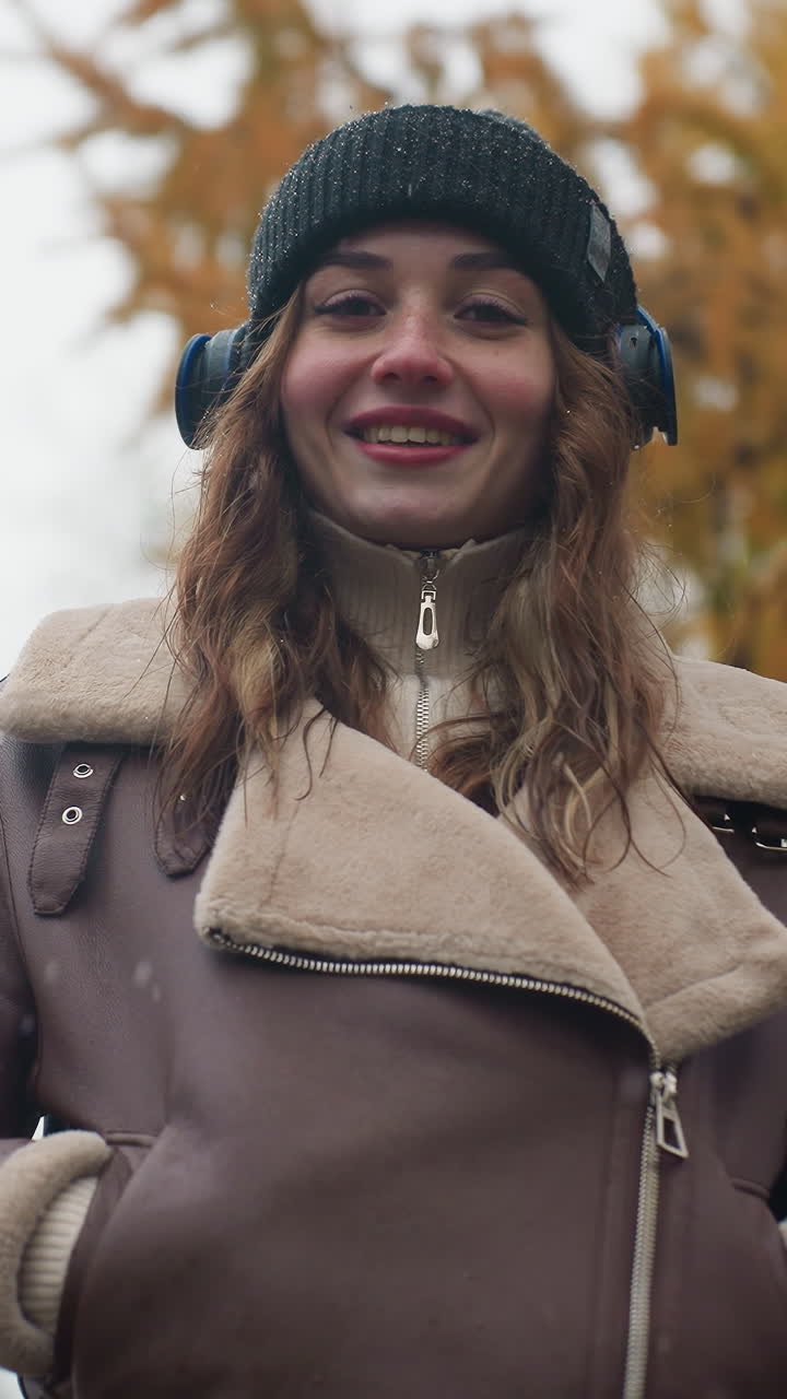 Smiling lady wearing black knit cap and brown shearling jacket with hands in pockets outdoors during cool autumn day, enjoying nature and fresh air