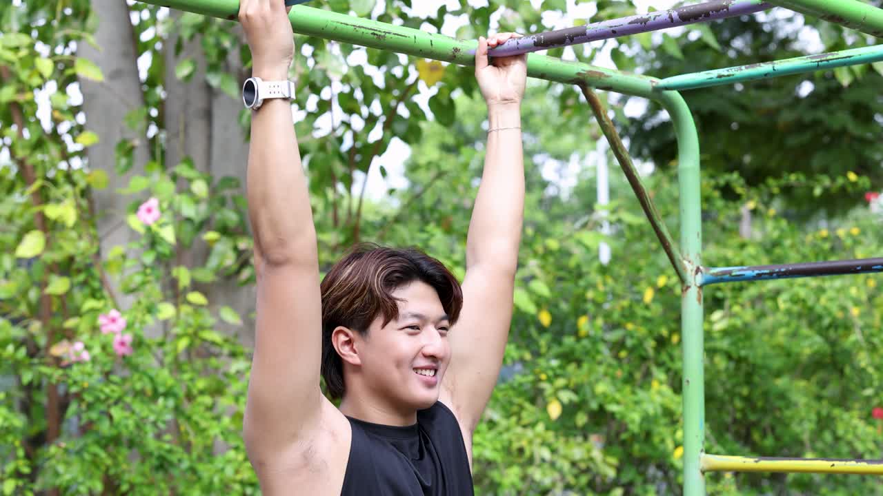 A young Asian man hangs from a green pull-up bar during an outdoor park workout