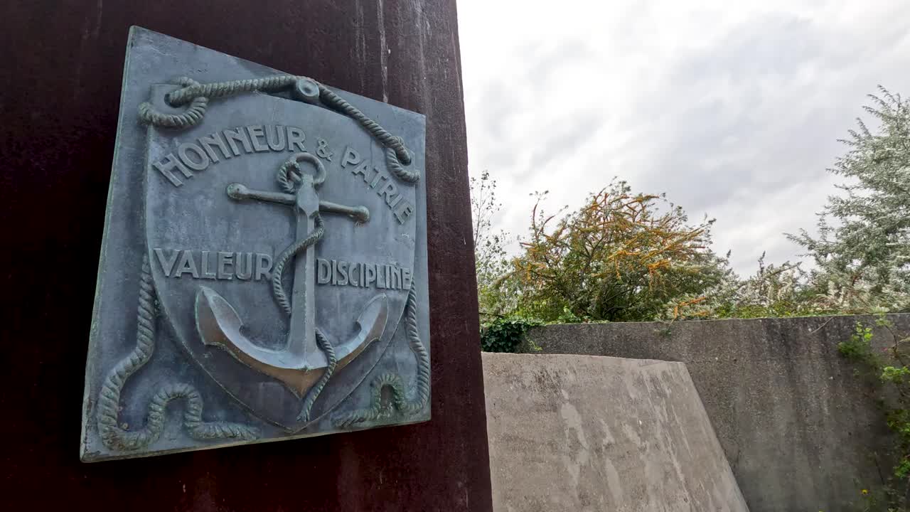 Camera slowly pans from a bronze naval plaque inscribed with 'Honneur Valeur Discipline' to an outdoor concrete memorial wall, under natural daylight with overcast skies
