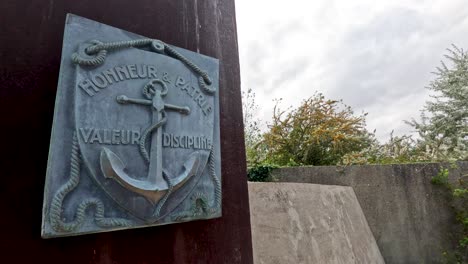 Camera slowly pans from a bronze naval plaque inscribed with &#039;Honneur Valeur Discipline&#039; to an outdoor concrete memorial wall, under natural daylight with overcast skies