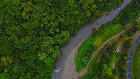 Drone-view-following-the-meandering-path-of-a-vibrant-jungle-river-with-a-road-next-to-it-before-panning-up-to-reveal-the-tropical-range-of-mountains-in-the-distance