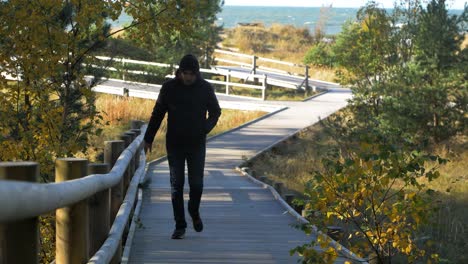 View-of-caucasian-male-exploring-nordic-seaside-forest,-wooden-pathway,-man-walking-alone-in-the-coastal-pine-forest,-sunny-day,-healthy-activity-concept,-medium-shot