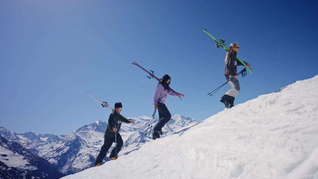 Inquadratura cinematografica di tre sciatori che risalgono una collina nella neve mentre trasportano gli sci a Verbier, Svizzera. L'iconico Grand Combin domina lo sfondo mentre il gruppo sale attraverso le Alpi svizzere.