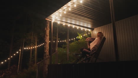 woman wraps in blanket sitting on folding chair. lonely woman enjoys silence of nature spending time on terrace of house decorated with light bulbs