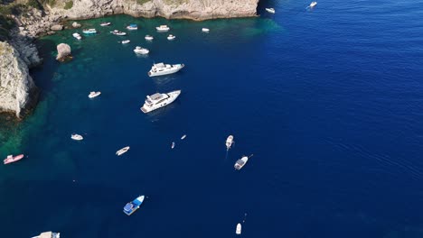 Boats-and-yachts-float-on-clear-blue-water-near-rocky-coastline